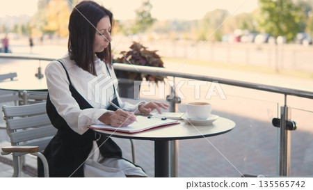 A professional woman sits at a round table outdoors, writing in a notebook. She enjoys a cup of coffee as she focuses on her work. The sun shines brightly, and trees are visible in the background. 135565742