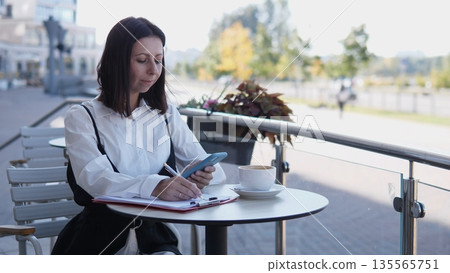 A business woman sits at a small table outside a cafe, focused on her phone and notes. She enjoys coffee while surrounded by a pleasant outdoor setting and warm sunlight. A business woman sits at a small table outside a cafe, focused on her phone and notes. She enjoys coffee while surrounded by a pleasant outdoor setting and warm sunlight. 135565751