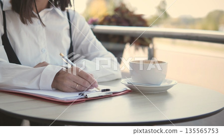 A business woman sits at a cafe table, writing notes on a clipboard while enjoying a cup of coffee. It is daytime, and the atmosphere feels relaxed and productive. A business woman sits at a cafe table, writing notes on a clipboard while enjoying a cup of coffee. It is daytime, and the atmosphere feels relaxed and productive. 135565753