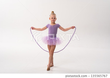 Young ballerina with a cochlear implant stretches a red hoop while posing in a lavender tutu. Inclusive gymnastics, hearing technology, and childhood coordination through rhythmic dance and movement. 135565767