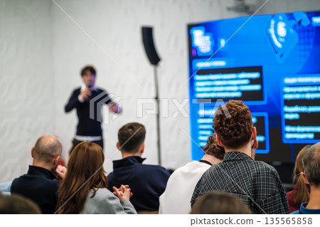Speaker presents to an audience in a modern conference room with a large blue screen Speaker presents to an audience in a modern conference room with a large blue screen 135565858