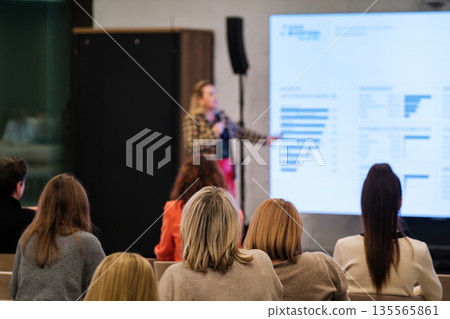 Speaker presents data to audience during conference with large screen charts and graphs. Speaker presents data to audience during conference with large screen charts and graphs. 135565861
