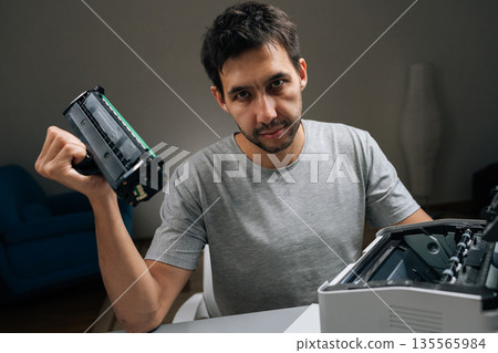 Young man holding empty toner cartridge from laser printer, looking directly at camera with frustrated expression while attempting to fix office equipment. Young man holding empty toner cartridge from laser printer, looking directly at camera with frustrated expression while attempting to fix office equipment. 135565984