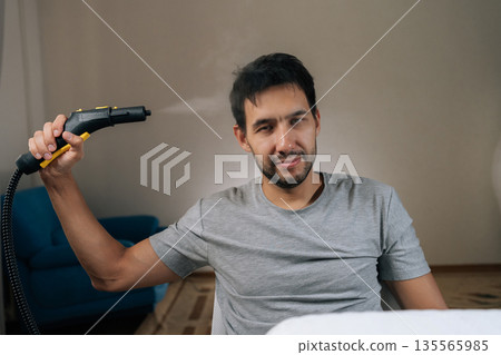 Portrait of young adult man playfully pointing steam cleaner upwards, releasing steam near head, embodying humorous and lighthearted approach to home cleaning tasks and daily routine. Portrait of young adult man playfully pointing steam cleaner upwards, releasing steam near head, embodying humorous and lighthearted approach to home cleaning tasks and daily routine. 135565985