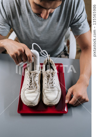 Top view of young adult man inserting portable electric dryer into white sneakers on red tray, demonstrating footwear care, moisture removal and hygienic drying to prevent odor and damage. Top view of young adult man inserting portable electric dryer into white sneakers on red tray, demonstrating footwear care, moisture removal and hygienic drying to prevent odor and damage. 135565986