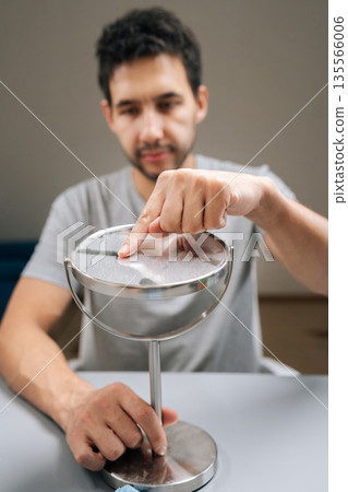 Man cleaning dusty vanity mirror, leaving distinct swipe mark with finger, highlighting effort in maintaining cleanliness and undertaking domestic tasks. Concept of domestic work, housekeeping. 135566006