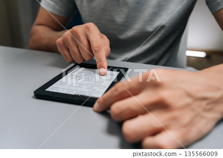 Close-up hands of unrecognizable male student using touchscreen e-reader siting at home desk, scrolling through e-book with finger, focused on portable device for study, leisure reading or research. 135566009