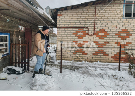 A man clears the snow from the yard. A man in a winter jacket and hat shovels snow from the yard of a rural farmhouse, clears the road and does winter chores outdoors on a cold frosty day. 135566522