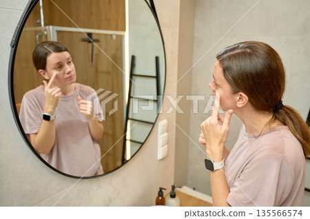 Woman standing in bathroom and touching face while looking at mirror Woman standing in bathroom and touching face while looking at mirror 135566574