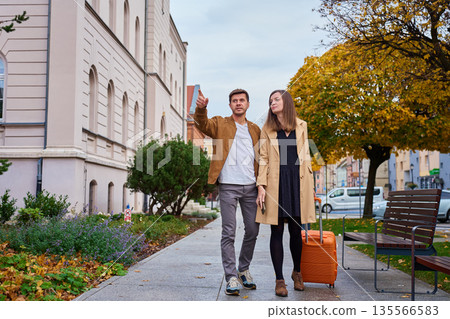 Man and woman walking on urban sidewalk with suitcase, man pointing direction 135566583
