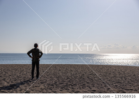 Silhouette of a man standing with his hands on his hips and looking out at the sea Silhouette of a man standing with his hands on his hips and looking out at the sea 135566916