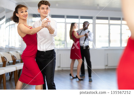 Positive woman in red dress and man practicing waltz dance moves in pair during group party in dancing studio Positive woman in red dress and man practicing waltz dance moves in pair during group party in dancing studio 135567252