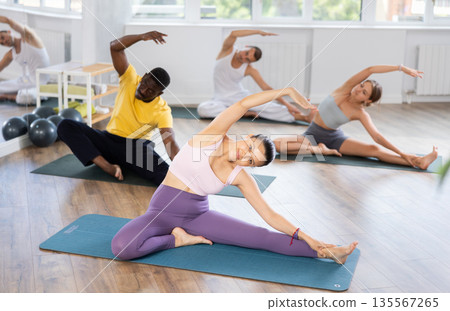 Women and men performing Parighasana side stretch asana, kneeling on leg on mat and extending arm overhead 135567265