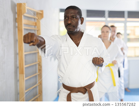 Middle-aged male attendee of karate classes practicing kata standing in row with others 135567282