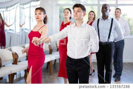 Young guy and woman in red dress waltzing during group class Young guy and woman in red dress waltzing during group class 135567286