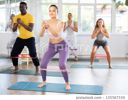 Asian girl with multinational friends training squats with difficulty, with elastic band on legs 135567330