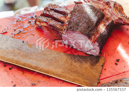 Cooked meat on a red cutting board with a wooden carving knife resting beside it, showcasing a well-seasoned crust and juicy interior during a barbecue preparation Cooked meat on a red cutting board with a wooden carving knife resting beside it, showcasing a well-seasoned crust and juicy interior during a barbecue preparation 135567337
