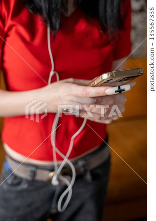 Young woman wearing a red shirt is holding a smartphone with earphones while sitting on a couch, casual indoor setting with soft lighting and relaxed atmosphere Young woman wearing a red shirt is holding a smartphone with earphones while sitting on a couch, casual indoor setting with soft lighting and relaxed atmosphere 135567338