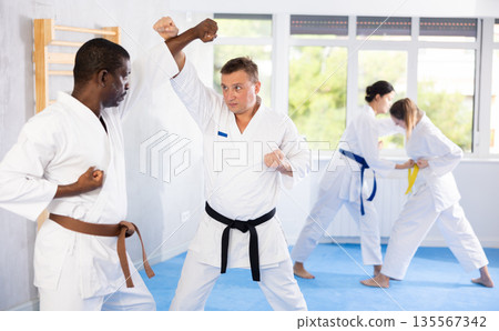 Middle-aged man attendee of karate classes fighting with his opponent in sports hall Middle-aged man attendee of karate classes fighting with his opponent in sports hall 135567342