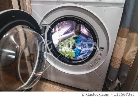 Front-loading washing machine with open door displaying colorful laundry inside, situated in a modern laundry room with tiled floor and curtains in the background Front-loading washing machine with open door displaying colorful laundry inside, situated in a modern laundry room with tiled floor and curtains in the background 135567355