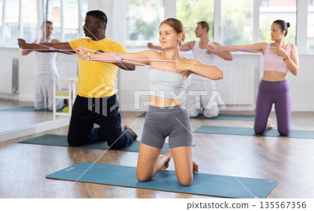 Young woman performs exercises with resistance band during multinational Pilates training, kneeling Young woman performs exercises with resistance band during multinational Pilates training, kneeling 135567356