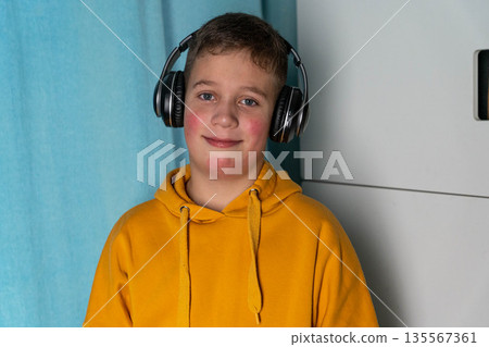 Young boy with headphones wearing a bright yellow hoodie is smiling in front of a light blue curtain, showcasing a casual indoor setting with a modern aesthetic Young boy with headphones wearing a bright yellow hoodie is smiling in front of a light blue curtain, showcasing a casual indoor setting with a modern aesthetic 135567361
