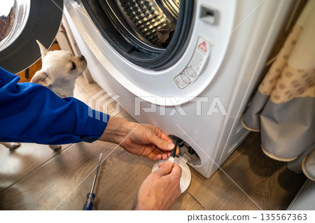 Person in blue shirt is repairing a washing machine while a small dog observes nearby in a home laundry room with wooden flooring and curtains in the background 135567363