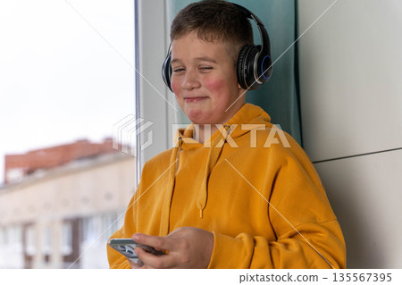 Young boy wearing headphones and a bright yellow hoodie is smiling while holding a smartphone near a window with a cityscape view in the background Young boy wearing headphones and a bright yellow hoodie is smiling while holding a smartphone near a window with a cityscape view in the background 135567395