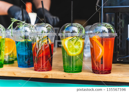 Colorful beverages in clear cups with fruit garnishes and herbs displayed on a wooden table at an outdoor event, showcasing refreshing drink options for summer gatherings 135567396