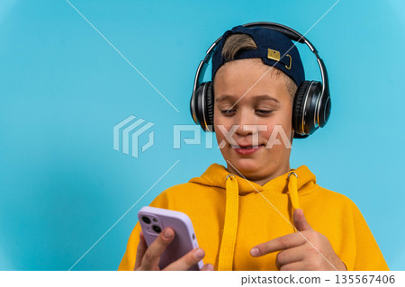 Young boy wearing headphones and a yellow hoodie is using a smartphone while smiling against a bright blue background with copy space for text or graphics 135567406