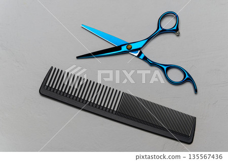 Blue scissors and black comb arranged on a gray surface, showcasing hairdressing tools for styling and grooming purposes in a professional setting Blue scissors and black comb arranged on a gray surface, showcasing hairdressing tools for styling and grooming purposes in a professional setting 135567436