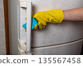 Hand wearing yellow rubber glove is holding a blue sponge while cleaning the handle of a white refrigerator in a kitchen setting with textured wall background 135567458