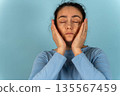 Woman with dark hair wearing a light blue long-sleeve shirt is holding her face with both hands, eyes closed, against a solid light blue background during a moment of reflection 135567459