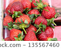 Fresh red strawberries with green leaves piled together in a clear container on a wooden surface, showcasing vibrant colors and natural textures for culinary use 135567460