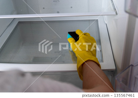 Person wearing yellow rubber gloves is cleaning the interior of a refrigerator with a sponge, showcasing a clear and organized space during a cleaning session 135567461