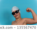 Young boy wearing a white swim cap and black goggles flexes his arm muscles against a bright blue background, showcasing confidence and readiness for swimming activities 135567470