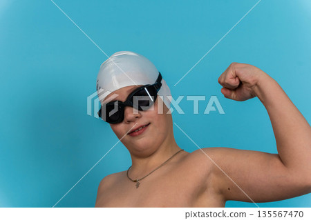 Young boy wearing a white swim cap and black goggles flexes his arm muscles against a bright blue background, showcasing confidence and readiness for swimming activities 135567470