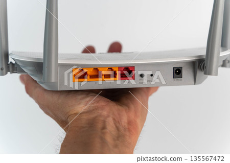 Hand holding a gray wireless router with visible Ethernet ports, power connection, and reset button, showcasing modern technology for home networking solutions Hand holding a gray wireless router with visible Ethernet ports, power connection, and reset button, showcasing modern technology for home networking solutions 135567472