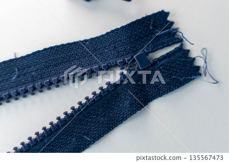 Close-up view of a broken navy zipper with frayed fabric edges on a light background, highlighting the details of the zipper teeth and pull tab for textile repair projects Close-up view of a broken navy zipper with frayed fabric edges on a light background, highlighting the details of the zipper teeth and pull tab for textile repair projects 135567473