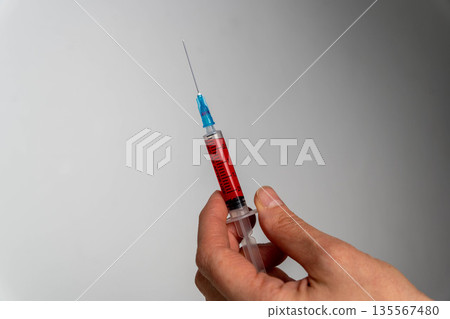 Hand holding a syringe filled with red liquid against a neutral background, showcasing the needle and plunger in a clinical context with copy space for text Hand holding a syringe filled with red liquid against a neutral background, showcasing the needle and plunger in a clinical context with copy space for text 135567480