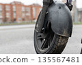 Close-up view of a black electric scooter wheel with a textured tire and mud splatters, parked on a city street with blurred buildings in the background 135567481