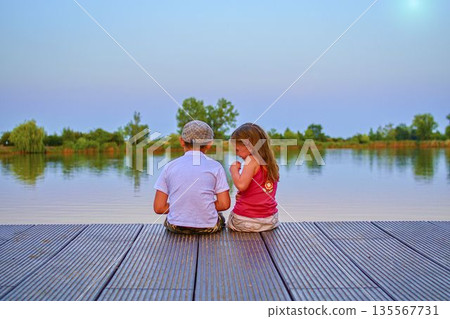 A little boy wearing a flat cap and a cute young girl sit together on a wooden pier by the lake. Small children sit on the pier at the side of a pond. The concept of a carefree childhood and summer A little boy wearing a flat cap and a cute young girl sit together on a wooden pier by the lake. Small children sit on the pier at the side of a pond. The concept of a carefree childhood and summer 135567731
