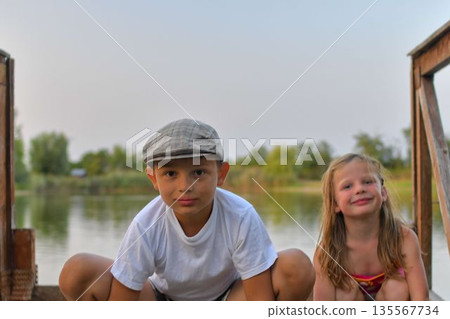 A little boy and cute girl kneel on a wooden pier on the shore of a lake. Small children sit on a pier at the edge of a pond. The concept of a carefree childhood and summer vacations. 135567734