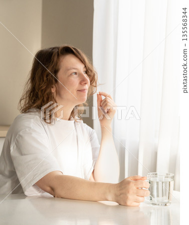 Woman takes pills with a glass of water at home in soft natural light. Daily medication and healthcare concepts 135568344