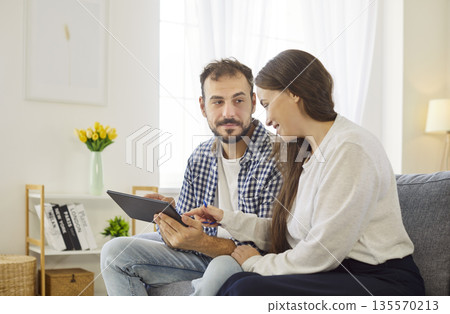 Couple using tablet with discussion at home, smiling wife pointing at screen Couple using tablet with discussion at home, smiling wife pointing at screen 135570213