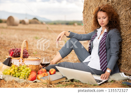 Woman in business suit spend time in field, settles down in nature with laptop and glass of wine Woman in business suit spend time in field, settles down in nature with laptop and glass of wine 135572327