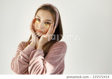Young woman applying a face patches. Brunette woman standing on a white backgroung. Girl wearing rose pajamas and headband. 135572414