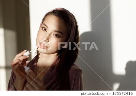 Young woman applying a lip gloss. Brunette woman standing in a bright studio. Girl wearing rose pajamas and headband. Young woman applying a lip gloss. Brunette woman standing in a bright studio. Girl wearing rose pajamas and headband. 135572416