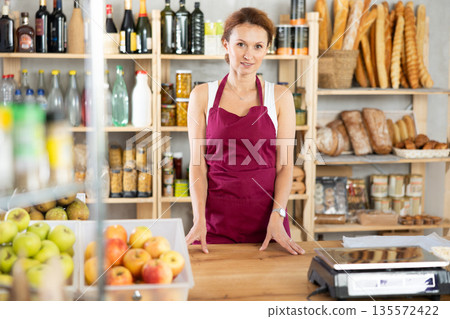 Portrait of positive female salesperson behind supermarket counter. Interior of grocery supermarket Portrait of positive female salesperson behind supermarket counter. Interior of grocery supermarket 135572422