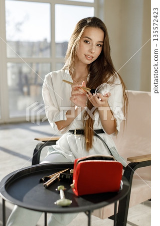 Young woman sitting on a chair and applying make up. Brunette woman sitting in a bright studio with big windows. Girl wearing white blouse and jeans. Young woman sitting on a chair and applying make up. Brunette woman sitting in a bright studio with big windows. Girl wearing white blouse and jeans. 135572423